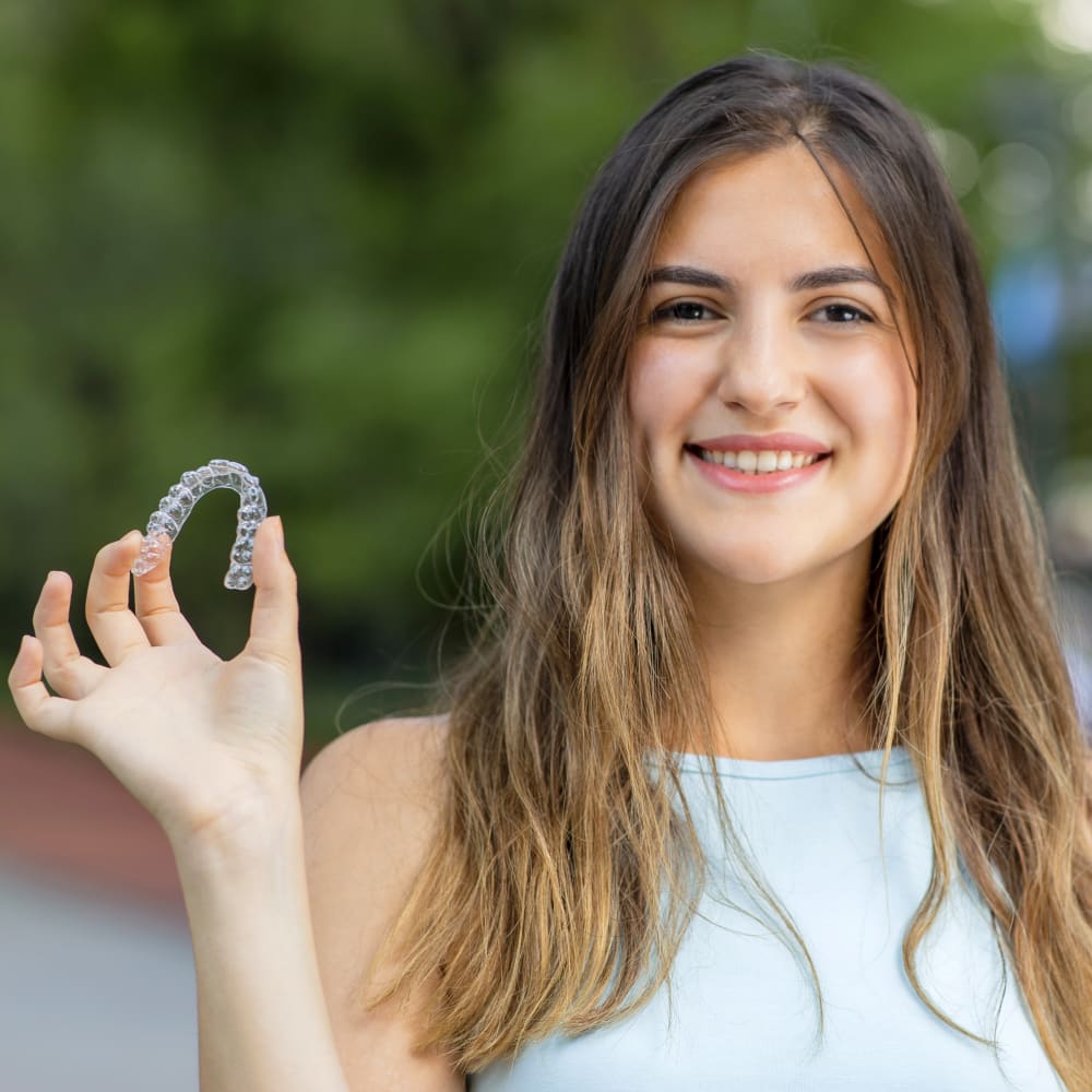 A young adult woman is smiling and holding up an Invisalign clear aligner.