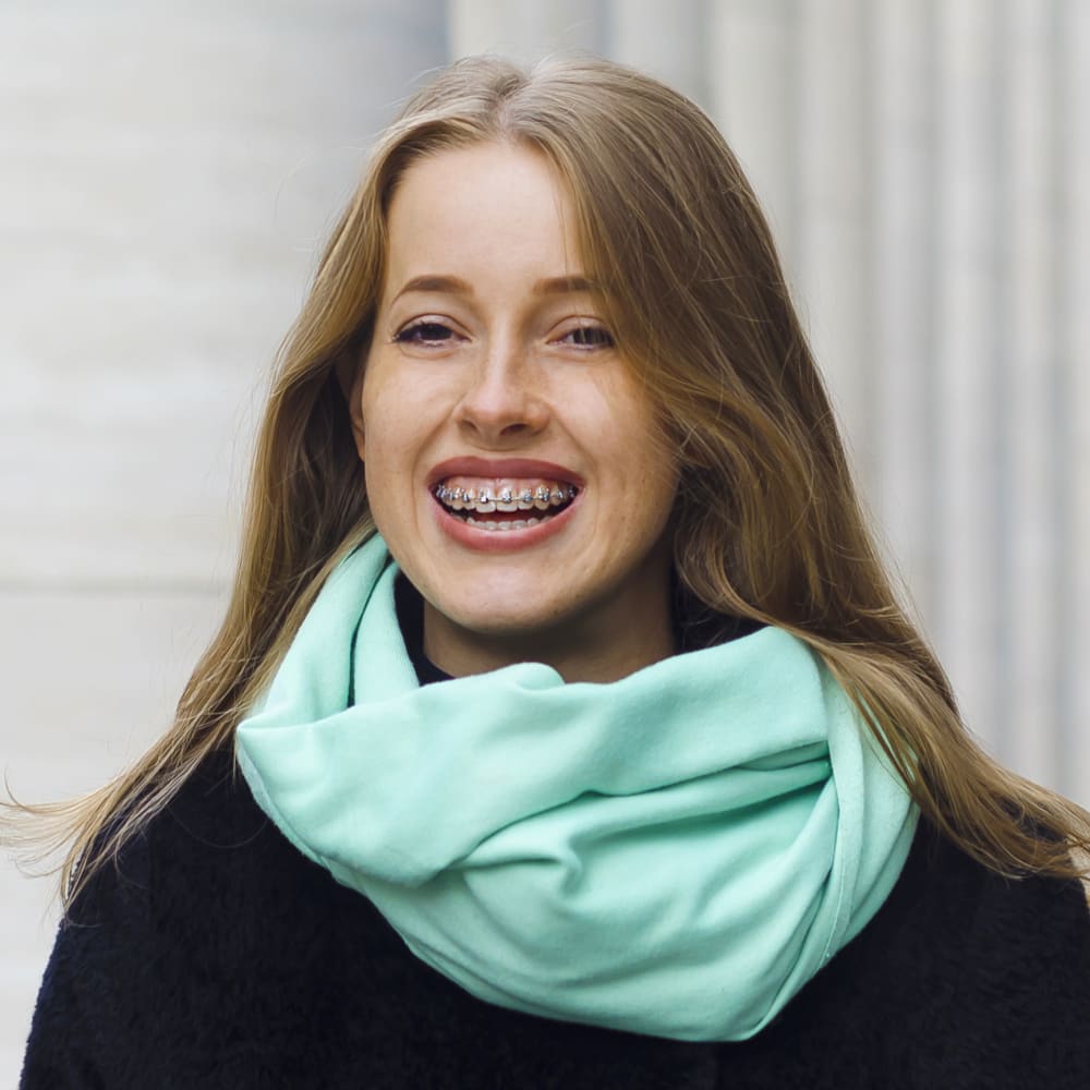 A woman with freckles wearing a seafoam green scarf is smiling, revealing a set of braces.