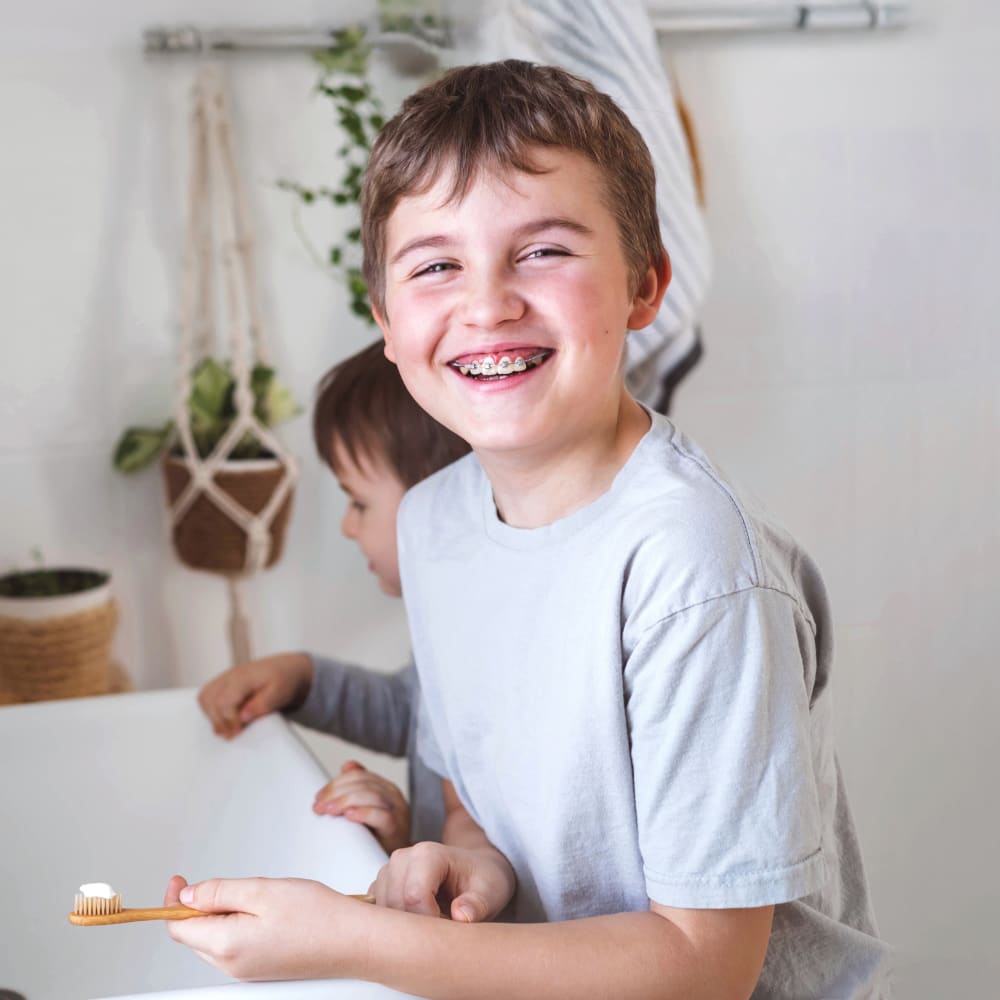 A young boy with braces is brushing his teeth at the sink with another young boy beside him.