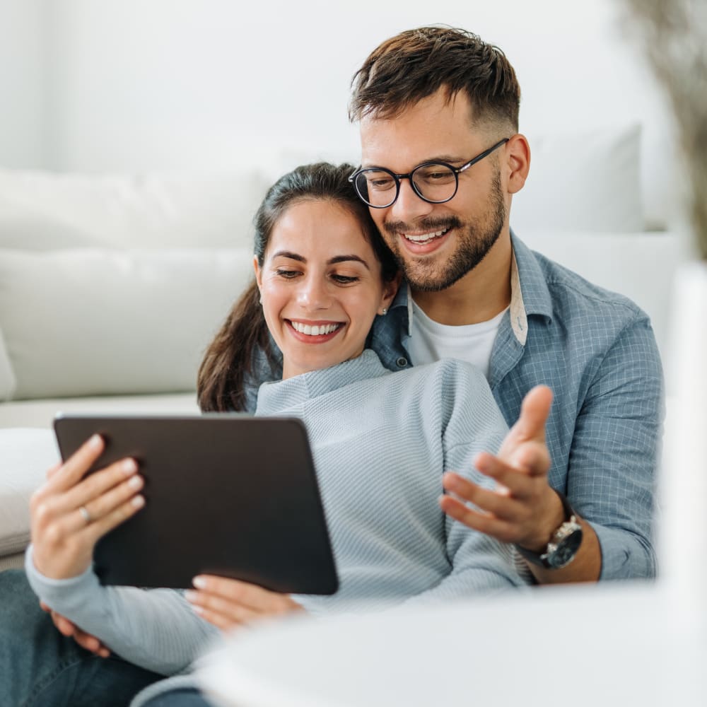 A woman is sitting in a mans lap with her back to him. They are both smiling at a tablet she is holding.