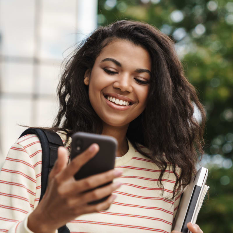 A teenage girl is smiling and looking at her smartphone. She is standing outside.
