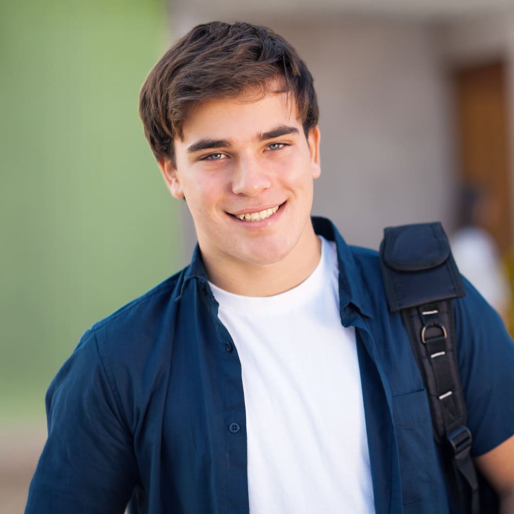 A teenage boy is smiling. He has a backpack slung over his left shoulder.