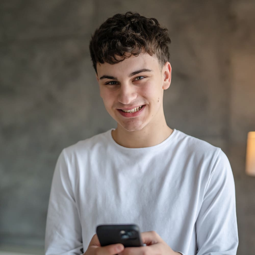 A teenage boy is smiling at the camera. He is also holding his smartphone.